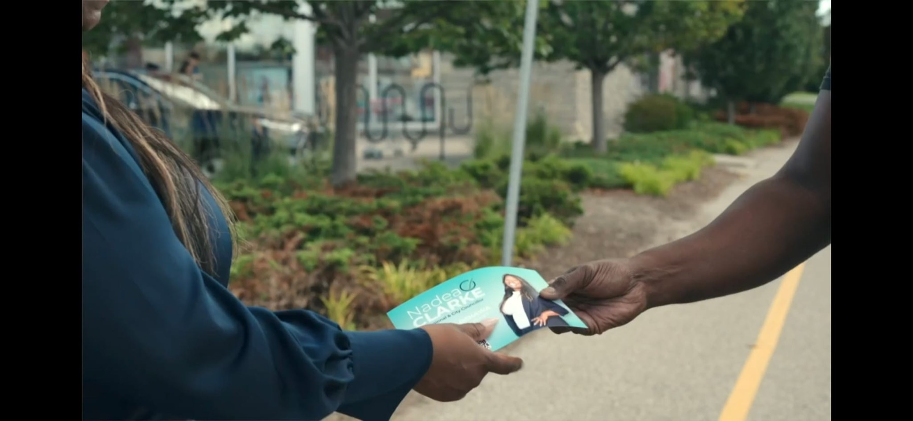A Nadea Clarke campaign card passing from one hand to another on a walkway in Ward 1.