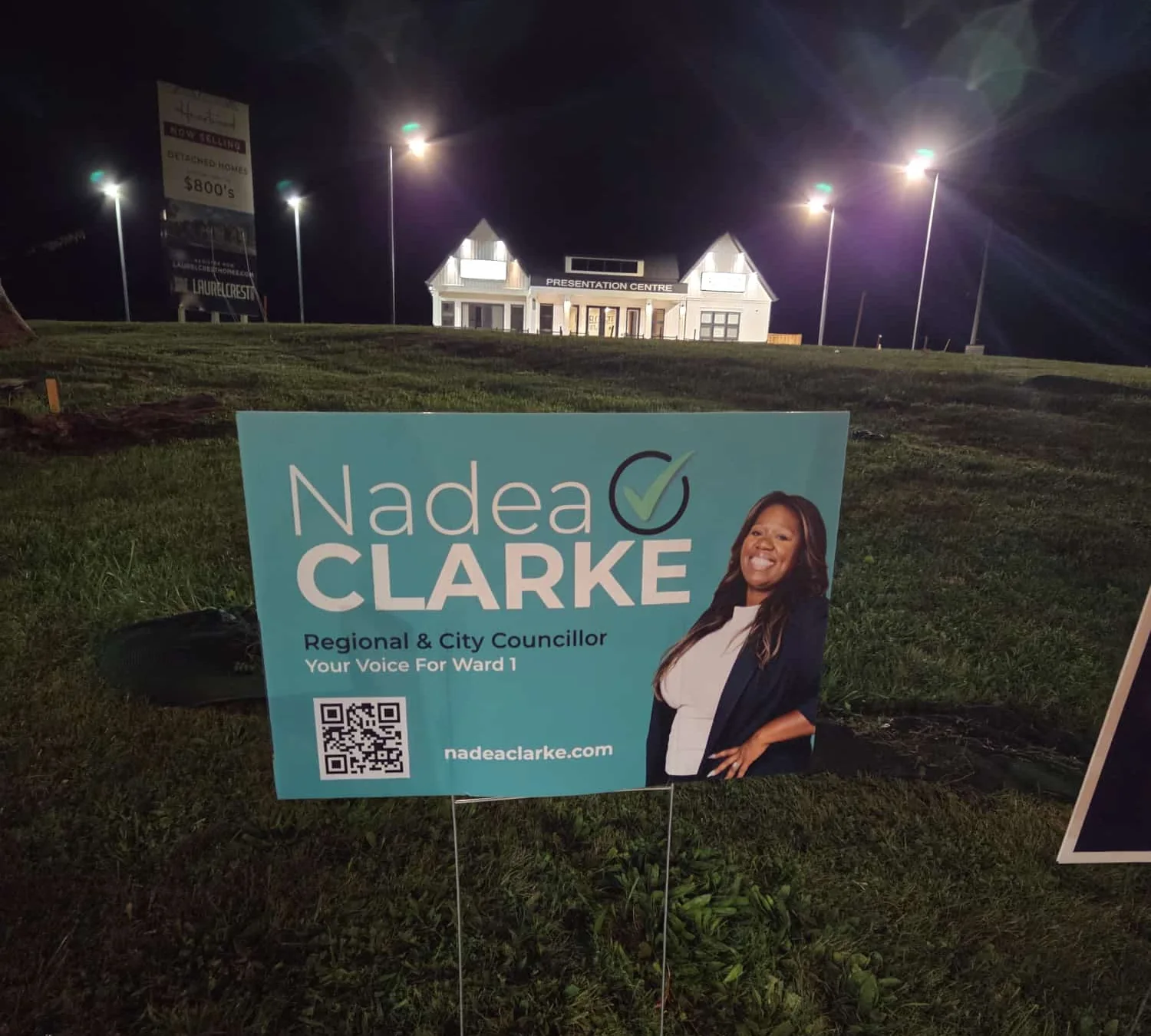 A Nadea Clarke campaign lawn sign — "Regional & City Councillor, Your Voice For Ward 1" — staked on grass at night outside a new-home presentation centre in Ward 1, Oshawa.